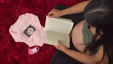 Eine-Schwangere-Frau-Sitzt-Gerne-Und-Liest-Ein-Buch,-Vor-Ihr-Liegen-Rosa-Babykleidung-Und-Ein-Ultraschallfoto