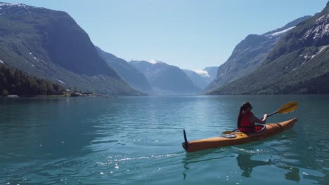Woman-kayaking-glacial-green-freshwater-lake-in-Loen---Beautiful-mountain-landscape-at-sunny-day---Camera-moving-sideways-close-to-water-with-kayak-moving-away-into-center-of-frame---Norway