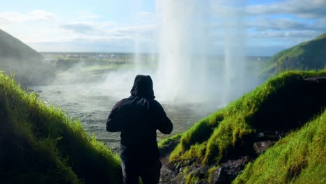 Male-Tourist-Wearing-Hooded-Jacket-With-Scenic-Waterfall-Of-Seljalandsfoss-In-Southern-Iceland