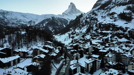 Aerial-flyover-over-Zermatt-city-center-during-winter-with-view-of-the-Matterhorn,-rooftops-and-river