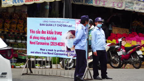 Static-view-of-policemen-talking-in-front-of-poster-related-to-Covid-rules-in-street-near-at-Da-Lat-market