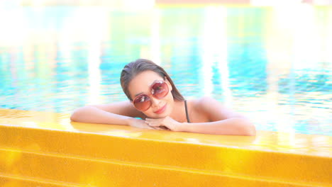 Beautiful-woman-in-red-sunglasses-resting-inside-swimming-pool-leaning-on-arms-at-the-pool-side-edge-at-exotic-Hotel-in-Bali