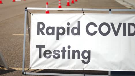 Rapid-Covid-19-Testing-site-sign-posted-outside-on-sunny-and-hot-summer-or-spring-day-in-empty-parking-lot-with-orange-cones-setup