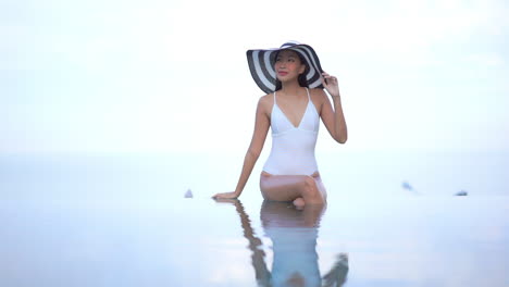 High-key-shot-of-beautiful-young-Asian-woman-with-big-hat-sitting-in-infinity-pool-edge