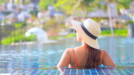 Exotic-woman-with-summer-hat-enjoying-in-infinity-pool-water-with-splendid-view-on-small-tropical-resort-city,-slow-motion-full-frame