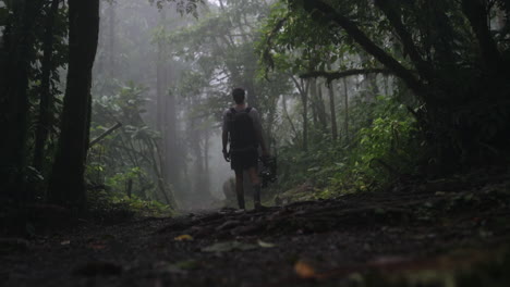 Man-walks-with-camera-equipment-down-foggy-rain-forest-path,-slow-motion