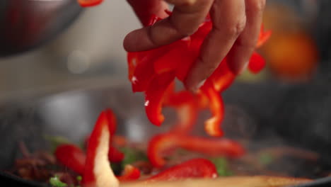 Hand-throwing-chopped-red-bell-peppers-into-a-frying-pan-with-cooked-onions