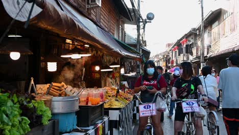 Zwei-Damen-Auf-Ihren-Fahrrädern-Halten-An,-Um-Thailändisches-Essen-Entlang-Der-Fußgängerzone-Zu-Kaufen,-Chiang-Khan-In-Loei,-Thailand