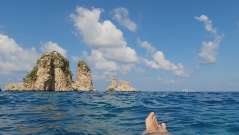 Personal-low-angle-perspective-of-legs-and-feet-floating-on-sea-water-with-Scopello-Stacks-or-Faraglioni-in-background