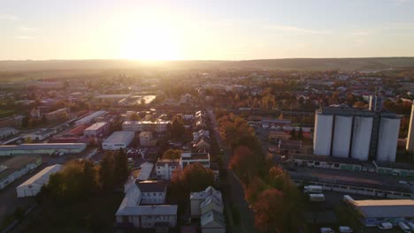 Drone-flight-over-autumn-street-with-trees-and-factory-complex-with-silo-at-sunset