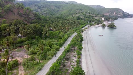 Aerial-rising-above-a-motorbike-travelling-along-a-rough-bumpy-gravel-coastal-road-surrounded-by-green-trees-next-to-the-ocean-on-idyllic-tropical-Atauro-Island,-Timor-Leste,-South-East-Asia