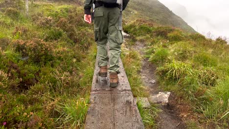Slow-motion-rear-view-of-hiker-with-dog-walking-on-narrow-wooden-trail-in-high-greened-mountains-of-Ireland---Rainy-day-with-dense-fog-in-the-valley
