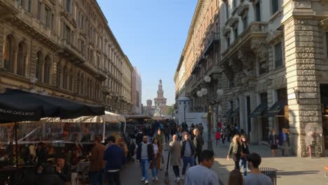 Via-Dante-pedestrian-street-in-Milan-with-people-strolling-and-shopping-with-Sforza-Castle-in-background,-Italy