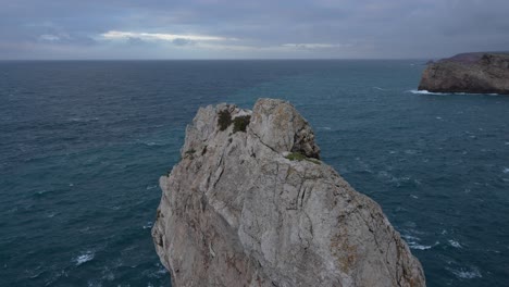 Roca-Gris-Que-Sobresale-Del-Agua-Azul-Del-Océano-Atlántico-En-El-Extremo-Suroeste-De-Portugal,-Durante-Una-Puesta-De-Sol-Nublada