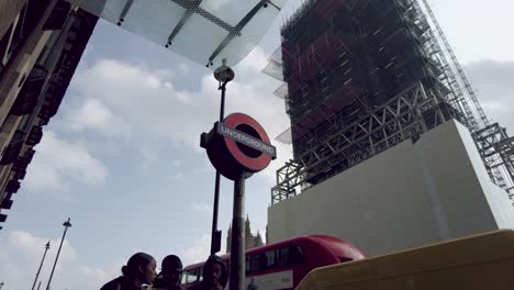 Upward-shot-as-pedestrians-walk-past-an-underground-sign-at-Westminster-station,-across-the-street-the-iconic-Big-Ben-completely-covered-in-scaffolding-as-it-undergoes-a-restoration,-London,-England