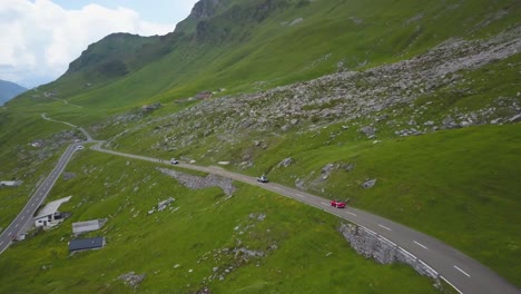 Aerial-dolly-in-of-vehicles-driving-on-a-paved-road-in-green-Klausen-Pass-mountainside-near-small-cottages-at-daytime,-Switzerland-Alps