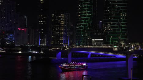 Puerto-Deportivo-De-Dubai-Por-La-Noche,-Barco-Iluminado-Con-Luces-De-Neón-Navegando-En-El-Canal-Bajo-Rascacielos