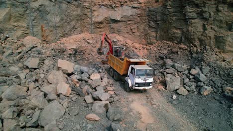Aerial-drone-view-of-Industrial-Excavation-Rotary-Truck-at-work-collecting-large-heavy-stones,-in-a-grey-mountain-clearing-with-labourers-looking-on-in-Vadodara,-India