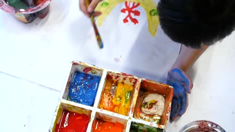 Overhead-shot-of-child-dipping-paintbrush-into-paint-pallet-to-get-blue-paint-for-painting-cute-designs-onto-large-sheet-of-white-paper-filmed-in-slow-motion
