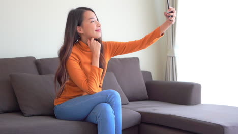A-pretty-young-woman-in-an-orange-sweater-and-blue-jeans-sits-on-the-couch-as-she-takes-several-selfies