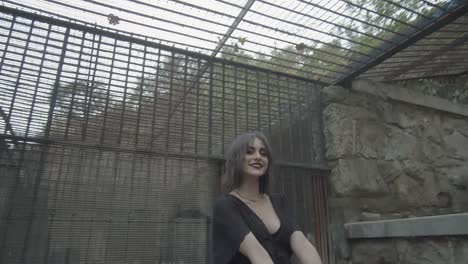 Woman-with-expressive-make-up-and-black-dress-stares-amusedly-at-the-sky-through-the-bars-of-the-cage-in-which-she-is-placed