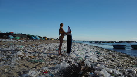 Caucasian-young-athletic-female-surfer-stand-in-polluted-beach-with-plastic-waste-trash-garbage,-Mother-Earth-ocean-pollution-save-the-planet-concept