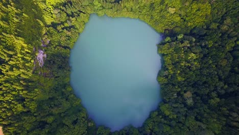 Drone-descent-towards-round-lake-of-turquoise-blue-water-surrounded-by-tropical-rainforest-on-a-volcanic-island