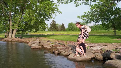 A-zoom-in-shot-of-two-child-sitting-on-a-rock-at-the-bank-of-a-lake-and-their-mother-keep-monitoring-standing-behind-them