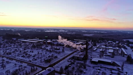 Aerial-forwarding-shot-over-urban-landscape-with-snow-covered-roofs-and-smoke-coming-out-of-an-industrial-chimney-at-sunrise