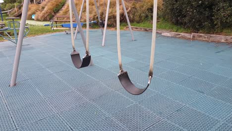 Kid's-empty-swings-swaying-and-swinging-in-the-breeze-in-empty-playground-with-safety-matting,-close-up-of-swings