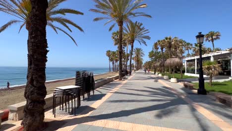 Walking-through-a-tropical-path-by-the-sea-on-a-sunny-day-with-blue-sky-and-palm-trees-in-Marbella-Spain,-holiday-destination-promenade-San-Pedro-de-Alcantara-nature,-4K-shot