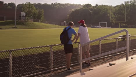 Runner-goes-up-to-coach-to-talk-on-bleachers-in-front-of-track-and-field-during-golden-hour,-runners-stretching-in-background