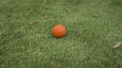Zeitlupe-Nahaufnahme-Eines-Orangefarbenen-Golfballs-Auf-Kunstrasen-Mit-Herumfliegenden-Blättern
