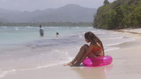 Latina-Girl-Enjoys-Playing-with-Sand-and-Water-Sitting-on-Tropical-Island-Beach-in-Inflatable-Ring