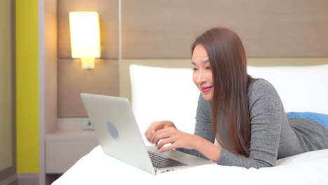 Vista-En-Cámara-Lenta-De-Una-Atractiva-Mujer-Asiática-Usando-Una-Laptop-Mientras-Está-Acostada-En-La-Cama-Bombea-Puños-Y-Gritos