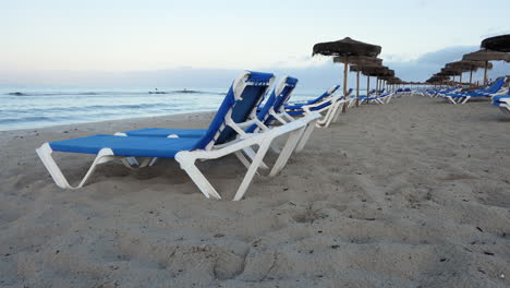 Side-view-of-an-empty-beach-at-dawn-with-many-sunbeds-and-straw-umbrellas,-small-waves-on-the-sea,-dream-luxury-travel-inspiration