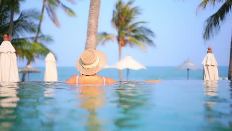 Back-of-Beautiful-Woman-in-Sunhat-Enjoying-the-View-from-Inside-Infinity-Pool-Leaning-on-the-Edge-at-a-Tropical-Island-Resort,-slow-motion