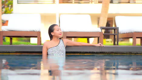 Young-asian-woman-rest-inside-water-on-the-edge-of-swimming-pool-next-to-line-of-deckchairs-at-sunset-Hotel-resort,-slowmotion