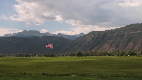 Gran-Bandera-Estadounidense-Ondeando-En-El-Viento-Sobre-Un-Campo-De-Hierba-Y-Un-Cielo-Nublado-Con-Las-Montañas-De-San-Juan-En-Segundo-Plano