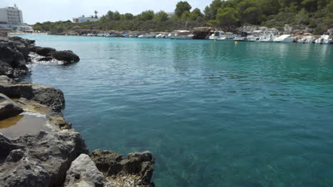 Malerischer-Blick-Vom-Wasserstand-Am-Mittags-Auf-Den-Bach-Cala-Santandria-Auf-Menorca-Mit-Gelben-Bootsmarkierungen,-Blauem-Transparentem-Meer-Und-Umliegenden-Felsen