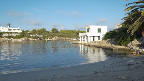 Seitenansicht-Bei-Sonnenaufgang-Des-Leeren-Strandes-An-Der-Bucht-Cala-Santandria-Auf-Menorca-Mit-Gelben-Bootsmarkierungen,-Blauem-Meer-Und-Umliegenden-Felsen