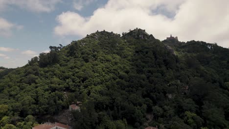 Aerial-ascending-shot-of-Sintra-hills-covered-with-lush-green-forest-and-mountaintop-castle-and-palace