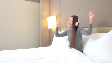 Young-woman-sitting-in-comfortable-bed-with-white-blanket-and-pillows-listening-and-dancing-to-music-playing-in-wireless-headphones