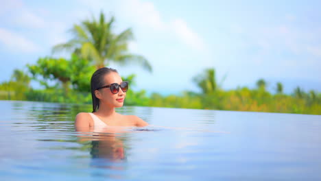Woman-touching-frame-of-sunglasses-inside-pool-water-in-a-tropical-paradise-on-sunny-day---slow-motion