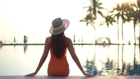 Rückansicht-Der-Frau-In-Einem-Orangefarbenen-Badeanzug,-Die-Bei-Sonnenuntergang-Am-Rand-Des-Swimmingpools-In-Einem-Exotischen-Hotel-Auf-Hawaii-Sitzt