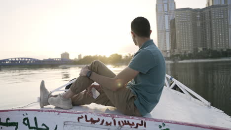 Push-in-as-man-sits-on-boat-with-Arabic-writing-watching-sunrise,-slow-motion
