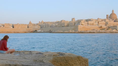 Frau-Sitzt-Auf-Felsen-Am-Meer-Mit-Blick-Auf-Die-Historischen-Gebäude-Von-Valletta-Im-Hintergrund-In-Malta