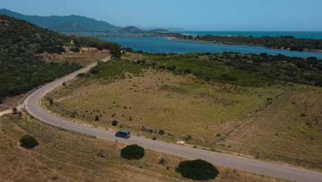 Aerial-of-VW-T4-Volkswagen-Bulli-hippie-van-car-is-driving-along-a-road-at-the-seaside-coast-of-the-island-Sardinia-in-Italy-with-the-blue-sea-landscape