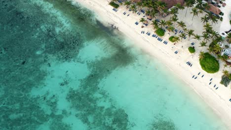Spinning-aerial-bird's-eye-drone-view-of-a-beautiful-tropical-vacation-beach-with-crystal-clear-blue-water,-white-sand,-palm-trees,-lounge-chairs-at-a-resort-in-Riviera-Maya,-Mexico-near-Cancun