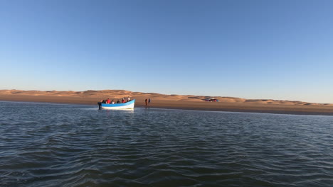 cinematic-shot-of-a-fishing-boat-that-moves-away-from-the-shore-of-the-Naila-lagoon,-the-dunes-and-another-boat-that-moves-away-can-be-seen